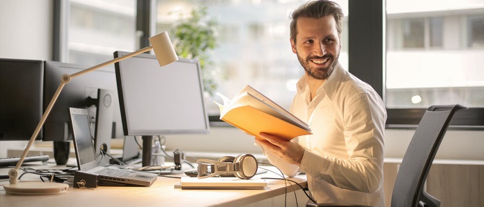 Man sitting behind his computer in an office with book in his hands