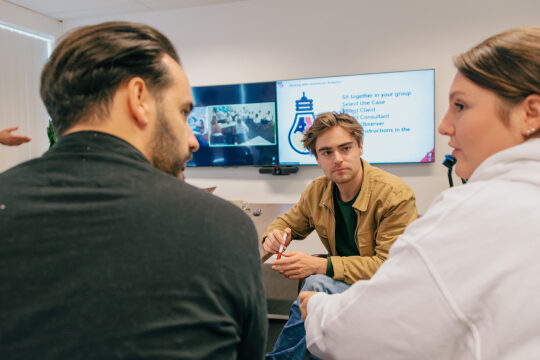 Three colleagues in discussion with flatscreen TVs in background